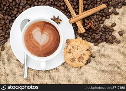 Cup of coffee with heart shape in foam with coffee beans and cinnamon sticks and star anise and cookie on hessian background