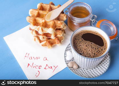 Cup of coffee with heart shaped sugar, homemade waffles with honey and a napkin with have a nice day message, on a blue wooden table.