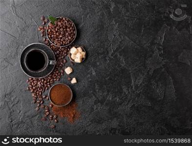 Cup of fresh raw organic coffee with beans and ground powder with cane sugar cubes with coffee tree leaf on dark background. Black ceramic mug. Space for text