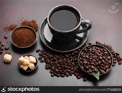 Cup of fresh raw organic coffee with beans and ground powder with cane sugar cubes with coffee tree leaf on dark background. Black ceramic mug