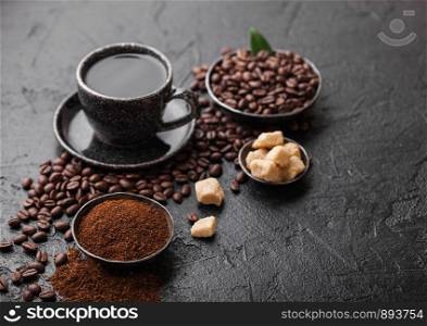 Cup of fresh raw organic coffee with beans and ground powder with cane sugar cubes with coffee tree leaf on dark background. Black ceramic mug.