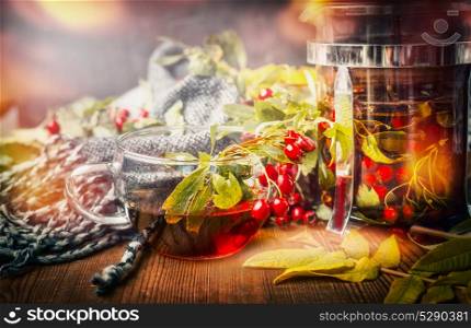 Cup of healthy tea with scarf, autumn leaves and berries on rustic wooden background. Hot autumn beverages concept