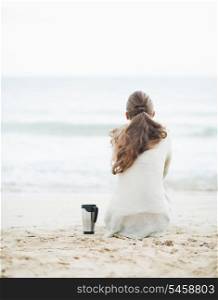 Cup of hot beverage near young woman in sweater sitting on lonely beach . rear view
