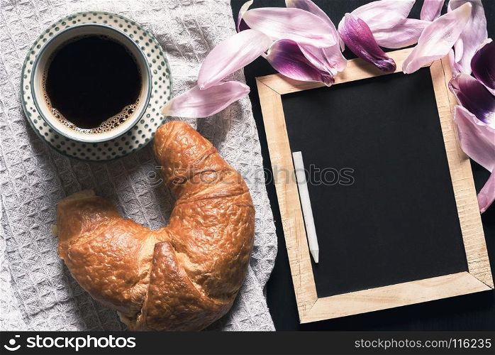 Cup of hot coffee and a fresh croissant on a retro kitchen towel, near a blank chalkboard, surrounded by pink tulip petals, on a black table.