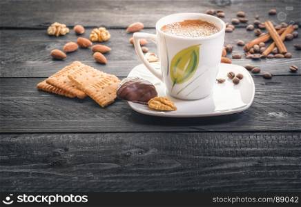 Cup of hot coffee on a plate, surrounded by tasty and healthy snacks, biscuits, almonds, walnuts and coffee beans, on an aged wooden table, in the morning light.