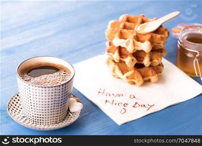 Cup of hot coffee with heart shaped sugar and tasty homemade waffles with honey. placed on a napkin with have a nice day message and a smiley face.