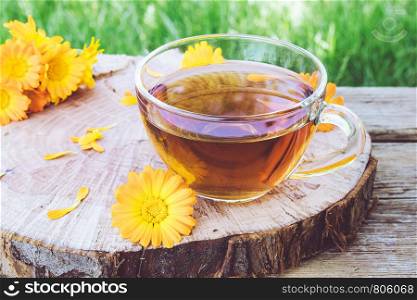 Cup of marigold and calendula tea flowers outdoors on green background. Extract of tincture of calendula in the cup. Medicinal plants.. Cup of marigold and calendula tea flowers outdoors on green background. Extract of tincture of calendula in the cup.