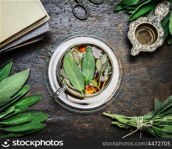 Cup of sage tea with fresh herbs leaves, books and old vintage strainer on rustic wooden background, top view, still life