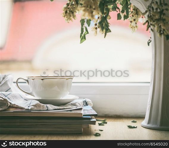 Cup on a stack of magazines at window with vase and flowers. Summer still life. Cozy home scene
