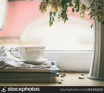 Cup on a stack of magazines at window with vase and flowers. Summer still life. Cozy home scene
