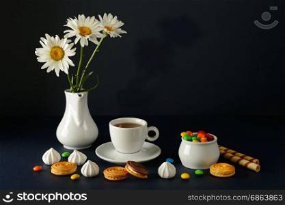 Cup with tea, teapot, sweets and white daisies on a black background