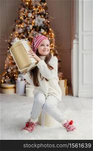 Curious small child doesn`t know what is in present box, recieves gift from Santa Claus, wears warm knitted clothes, sits against decorated New Year tree background. Childhood, surprisment, holidays