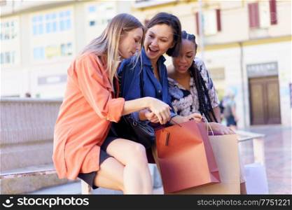 Curious young diverse ladies looking inside shopping bag of cheerful female friend while sitting together on city square. Positive young ladies examining purchases of surprised female friend