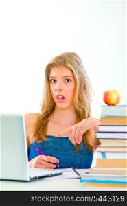 Curious young girl sitting at table with books and pointing on laptop &#xA;