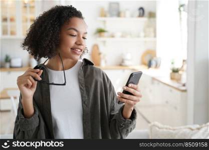 Curly african american girl has fun with phone at home. Young woman browsing social media and smiling. Chatting with friends remotely or watching video. Messaging with classmates and sharing content.. Curly african american girl has fun with phone. Messaging with classmates and sharing content.