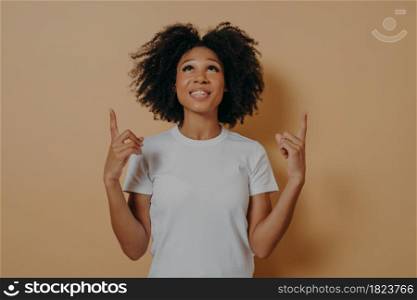 Curly african woman 20s pointing with forefingers upwards at copy space with smile on face, isolated over brown wall background, studio shot of dark skinned lady presenting or displaying product. Curly african woman pointing with forefingers upwards at copy space against brown studio wall