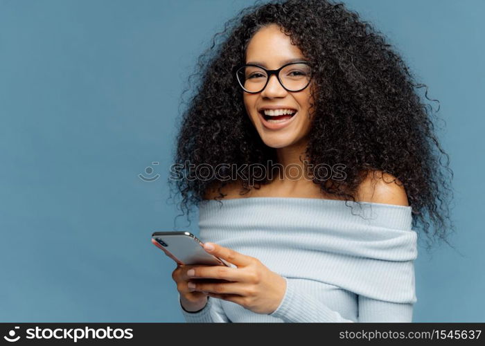 Curly cheerful woman with pleased expression, holds mobile phone, checks email box, wears optical glasses and jumper, enjoys using modern technologies, isolated over blue background, blank space