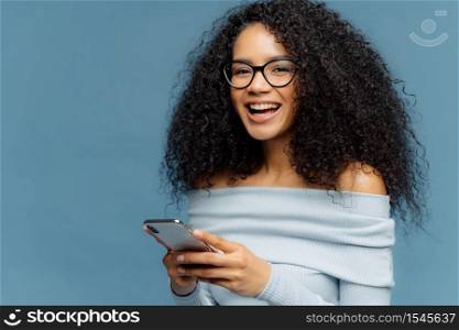 Curly cheerful woman with pleased expression, holds mobile phone, checks email box, wears optical glasses and jumper, enjoys using modern technologies, isolated over blue background, blank space