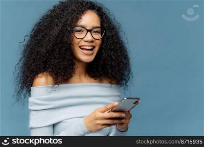 Curly cheerful woman with pleased expression, holds mobile phone, checks email box, wears optical glasses and jumper, enjoys using modern technologies, isolated over blue background, blank space