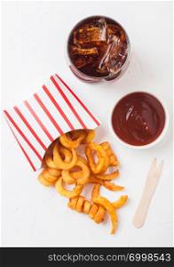 Curly fries fast food snack in paper container with glass of cola and ketchup on kitchen background. Unhealthy junk food