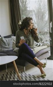 Curly hair young woman using mobile phone and holding mug while sitting on sofa in the room