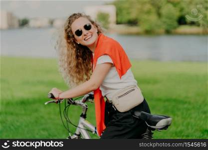 Curly haired blonde woman cycles outdoor, stands with bicycle, carries bag, wears sunglasses, poses against green lawn, wears white t shirt, black trousers, red sweater on shoulders. Outdoor lifestyle