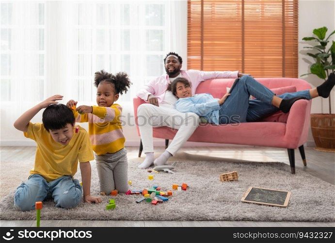 Curly little kindergarten girl teasing cute boy while playing wooden blocks together on floor in living room. Couple parent resting on couch while father scolding daugther and mother watching children.