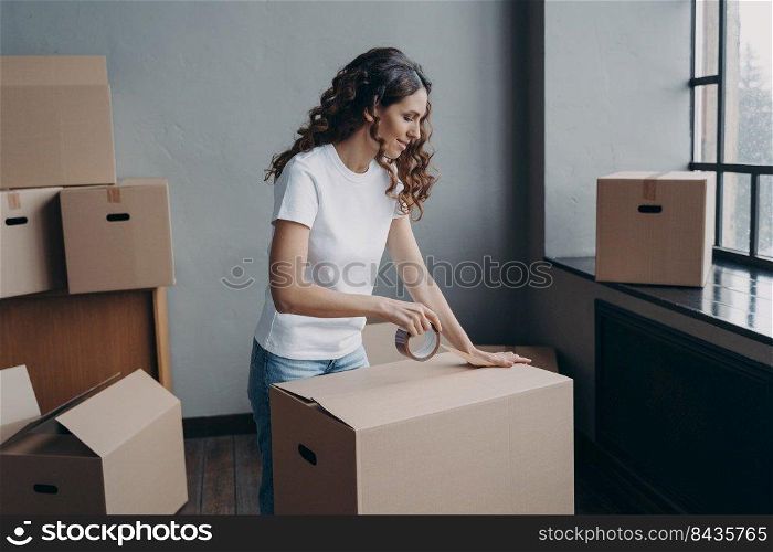 Curly spanish girl is ready for relocation. Lady is wrapping cardboard boxes with packing tape. Happy young woman purchasing or selling real estate and going to send things as cargo.. Curly spanish girl is ready for relocation. Lady is wrapping cardboard boxes with packing tape.