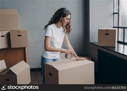 Curly spanish girl is ready for relocation. Lady is wrapping cardboard boxes with packing tape. Happy young woman purchasing or selling real estate and going to send things as cargo.. Curly spanish girl is ready for relocation. Lady is wrapping cardboard boxes with packing tape.