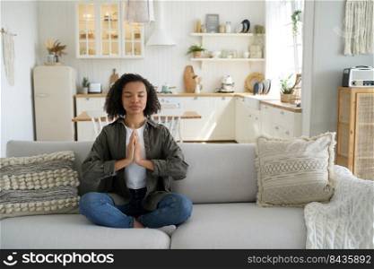Curly young hispanic woman is sitting on couch in lotus pose and meditating. Teenage afro girl is practicing yoga at kitchen at home. Concentration and balance. Wellness and stress relief.. Curly young woman sitting on couch in lotus pose and meditating. Wellness and stress relief.