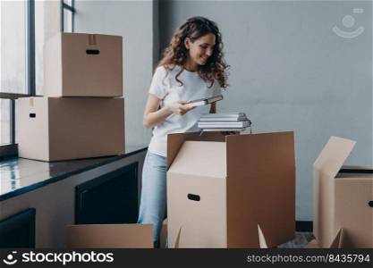 Curly young woman, student is housing in residential room. Happy european woman unpacking boxes with books. Girl in white t-shirt indoors. Relocation and independence concept.. Young woman, student is housing in residential room. European woman unpacking boxes with books.