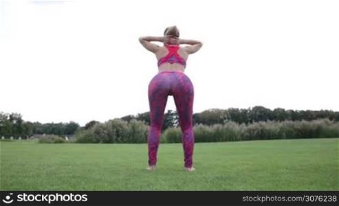 Curvy woman doing squats with hand behind her head in the park on green grass. Rear view of woman exercising outdoors on a nice summer day.