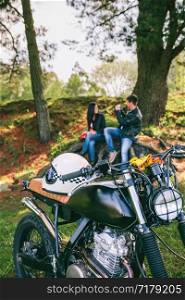Custom motorbike with young couple resting from a motorcycle trip in background. Selective focus on motorbike in foreground. Custom motorbike with young couple