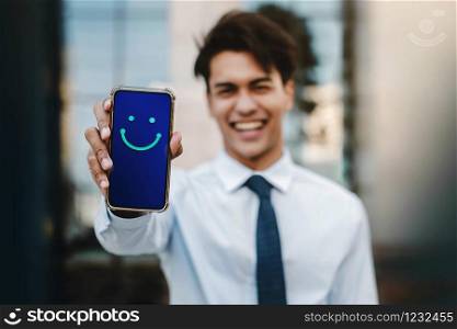 Customer Experiences Concept. Young Businessman Giving a Happy Face Icon and Positive Review via Smartphone. Client&rsquo;s Satisfaction Surveys on Mobile Phone. Front View