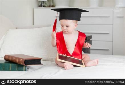 Cute 10 months old baby boy in graduation cap holding digital tablet and looking at stack of books