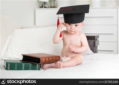 Cute 10 months old baby boy siting on bed with books and taking off graduation cap