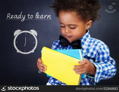 Cute african american boy with books in hands on blackboard background, adorable preschooler ready to learn in elementary school