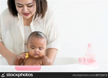 Cute African newborn baby bathing in bathtub with soap bubbles on head and body while playing rubber duck toy. Mother washing her little daughter in warm water. Newborn baby cleanliness care concept