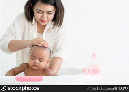 Cute African newborn baby bathing in bathtub with soap bubbles on head and body. Asian young mother washing her little daughter in warm water. Newborn baby cleanliness care concept