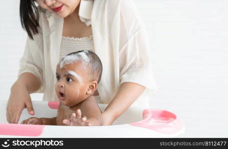 Cute African newborn baby bathing in bathtub with soap bubbles on head and body. Asian young mother washing her little daughter in warm water. Newborn baby cleanliness care concept