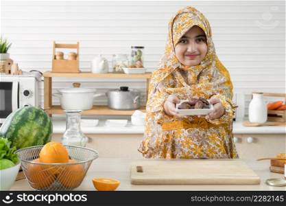 Cute Arab muslim kid girl wearing hijab smiling and holding a plate full of sweet dry dates fruit on iftar time in ramadan kareem in kitchen at home with oranges, watermelon, vegetables and milk
