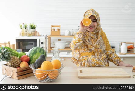 Cute Arab muslim kid girl wearing hijab smiling and holding a sliced orange cover one eye in kitchen at home with many fruits, vegetables and milk on table for doing healthy food