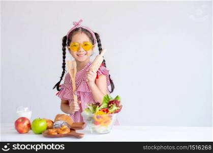 Cute Asian girl shows kitchenware with various types of fruit and vegetables and stand in front of white wall background.