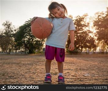 Cute Asian little girl in sportswear holding a basketball and looking at the camera at summer park. Healthy outdoor sport for young child.