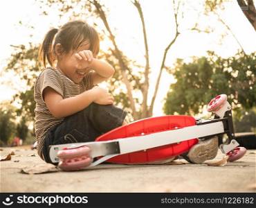 Cute Asian little girl sitting on the ground after falling off her scooter at summer park. Healthy outdoor sport for young child.