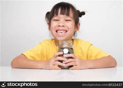 Cute Asian little girl smiling happily holding money coins in clear jar sitting at a table on white background and looking at the camera. Children learning about saving for future concept
