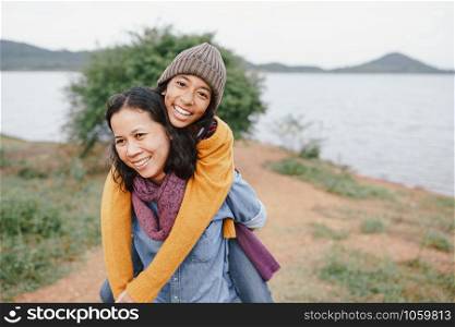 Cute Asian mother and daughter piggyback ride on her back with the laughing at river in the morning. The concept of family tourism holiday lifestyle.