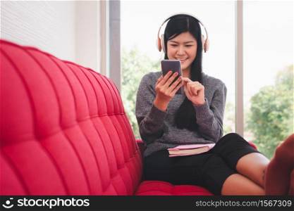 Cute Asian woman is sitting on the sofa in the morning And send messages on mobile phones While listening to music And with a book on the lap In the living room