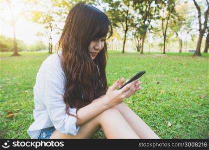 Cute asian woman reading pleasant text message on mobile phone while sitting in park spring day. Asian woman using on smart phone with feeling relax and smiley face. Lifestyle and technology concepts.