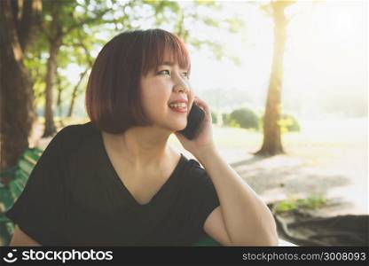 Cute asian woman smiles and talking on mobile phone while sitting in park spring day. Asian woman using on smart phone with feeling relax and smiley face. Lifestyle and technology concepts.